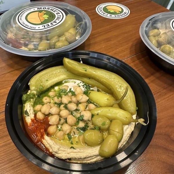 A black bowl of Moshe's Falafel hummus with chickpeas, green sauce, red sauce, and pickles.