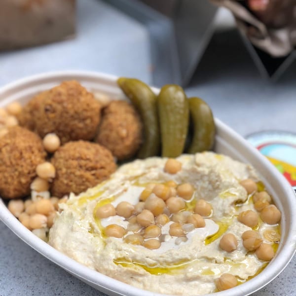 A white bowl of Moshe's falafel, hummus with chickpeas and olive oil, and pickles.