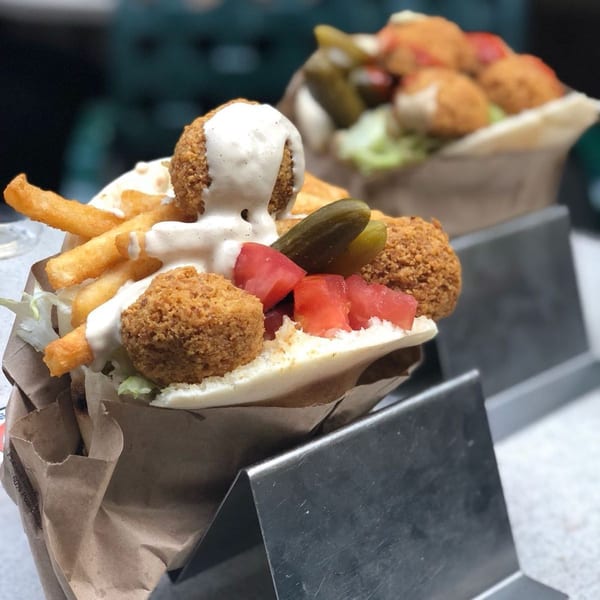 Two Moshe's falafel pita sandwiches with fries, pickles, tomatoes, and white sauce, in metal holders.