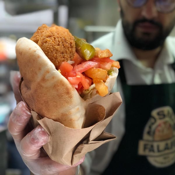 A gloved hand holds a Moshe's falafel pita sandwich filled with falafel, fries, tomatoes, and a pickle.