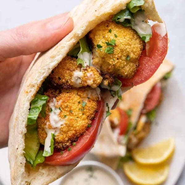 Hand holding a Moshe's falafel pita with crispy cauliflower, lettuce, tomato, and white sauce.