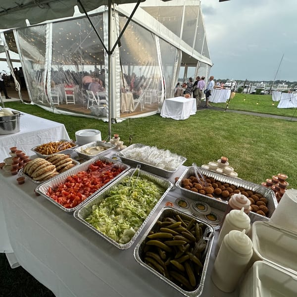 Outdoor buffet table with falafel, pita, chopped vegetables, and sauces, next to a clear event tent.